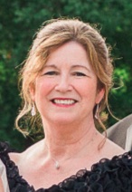 A close-up portrait of a smiling middle-aged woman with light brown hair styled in a soft updo, wearing dangle earrings, a subtle necklace, and a black ruffled outfit against a blurred green background